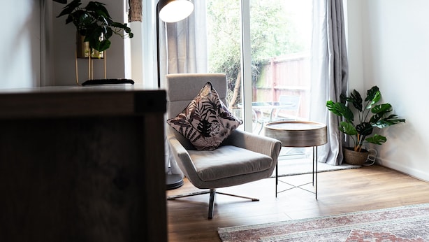A modern living room with a grey armchair, a round side table, a floor lamp, and a large window with curtains. - Inviting 4-bed House in Kettering (Kettering)