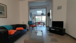 Sala de estar com sofá preto, mesa de centro e televisão. Ao fundo, uma área de jantar com cadeiras e mesa.