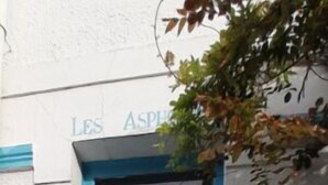 Uma porta azul com duas maçanetas, uma pequena janela acima e uma parede branca com uma placa sobre a porta.