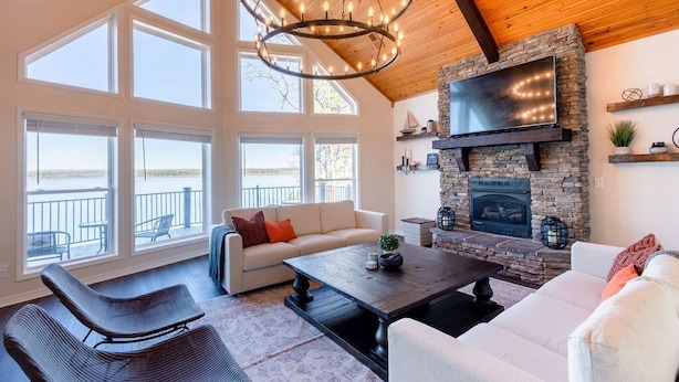 A spacious living room with a stone fireplace, large windows, and a wooden ceiling.