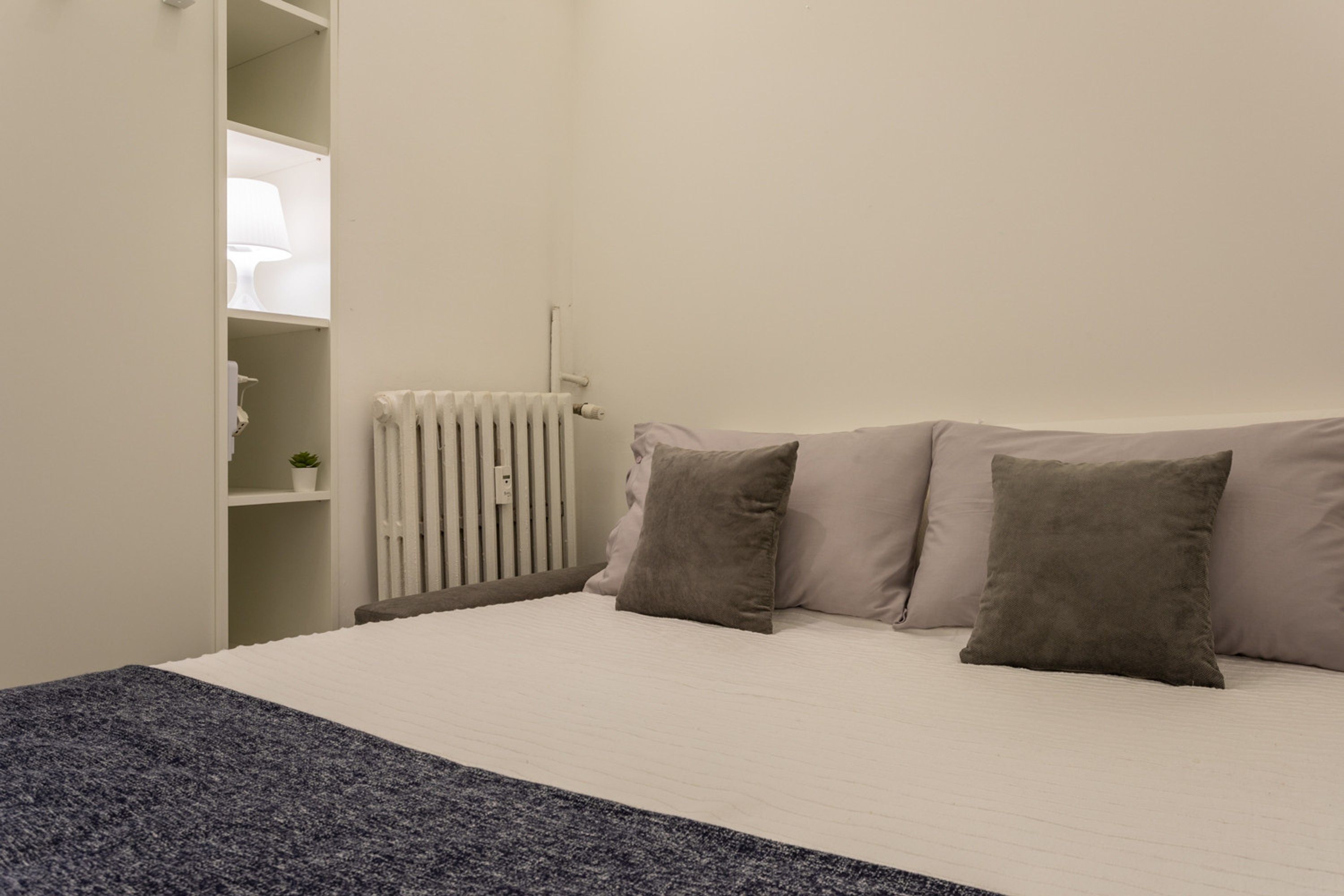 A bedroom with a bed, pillows, a radiator, a shelf with a lamp, and a small plant.