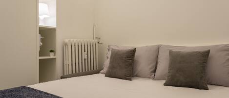 A bedroom with a bed, pillows, a radiator, a shelf with a lamp, and a small plant.