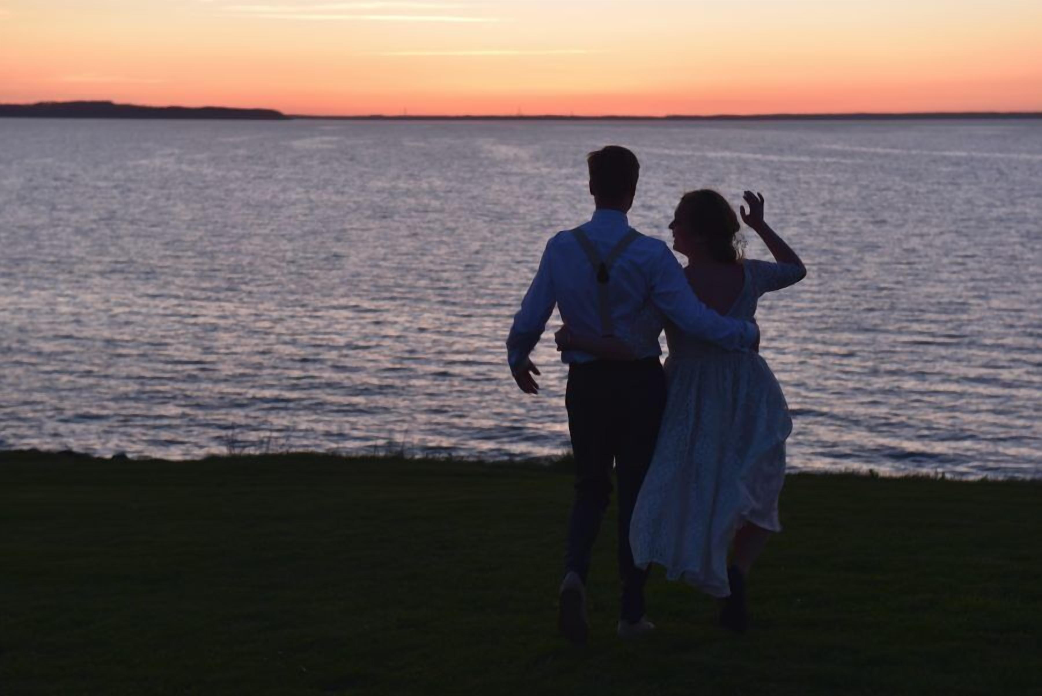 A couple standing on a grassy area facing a body of water at sunset.