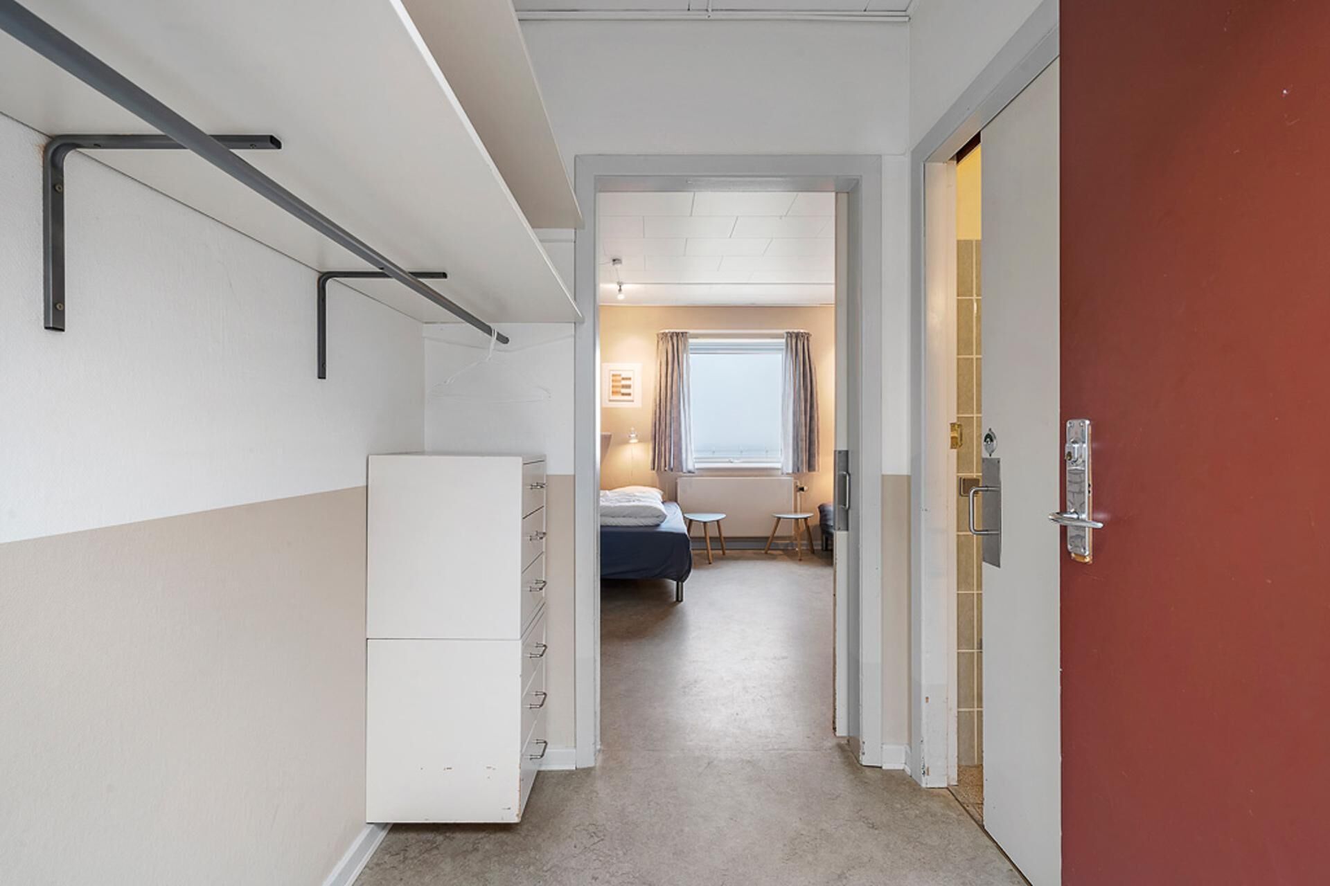 Ein schmaler Flur führt zu einem Schlafzimmer mit einem Bett, einem kleinen Tisch und einem Fenster. Rechts befinden sich ein weißer Schrank und eine rote Tür.