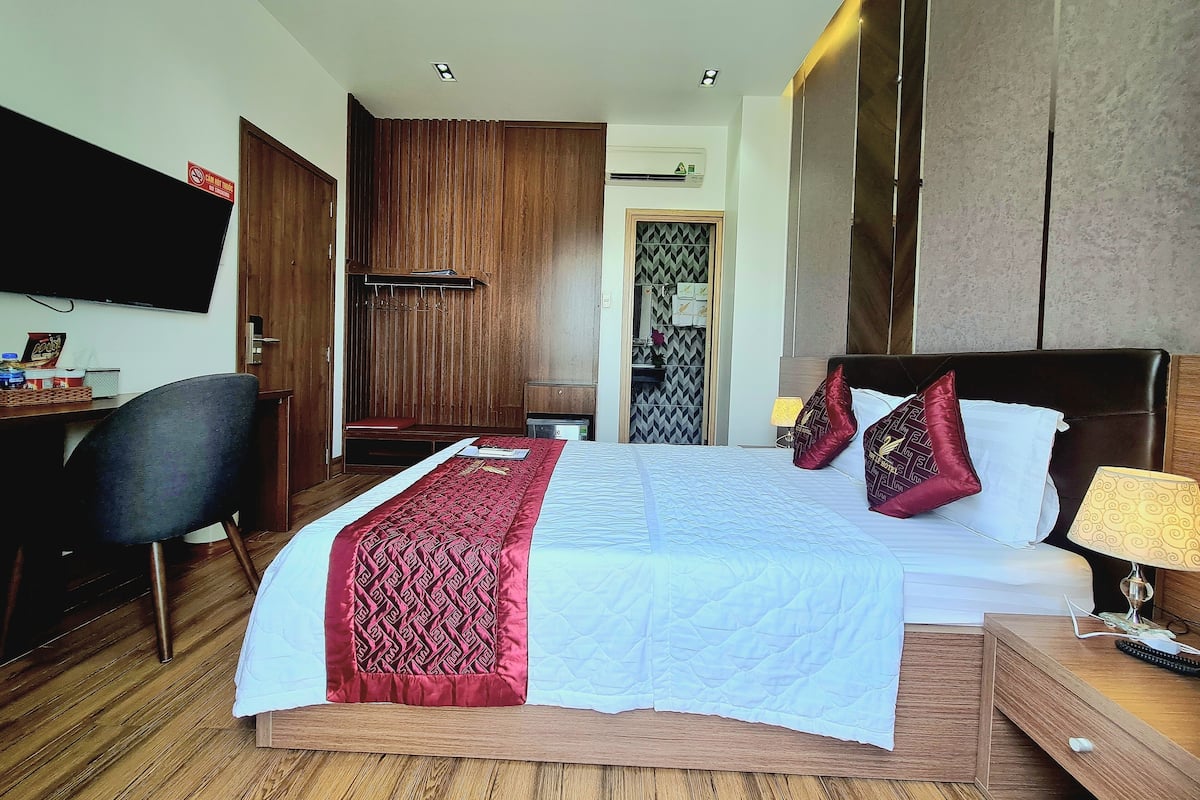 Quarto de hotel com cama, mesa de cabeceira, luminária e escrivaninha com cadeira.