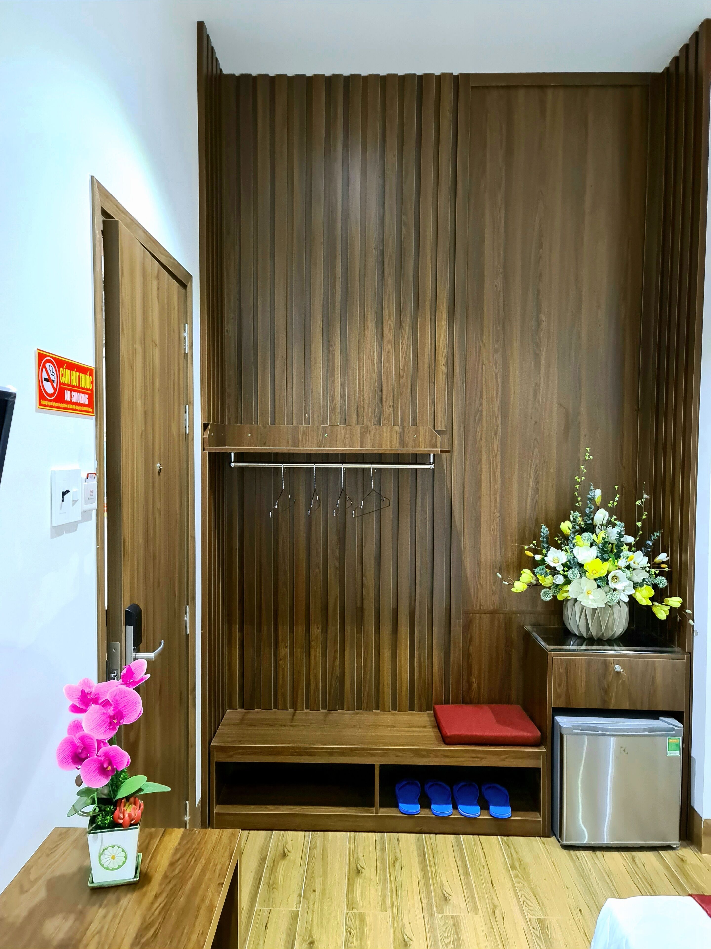 A room with a wooden wardrobe, a small table with a flower vase, a red cushion, and a metallic refrigerator.