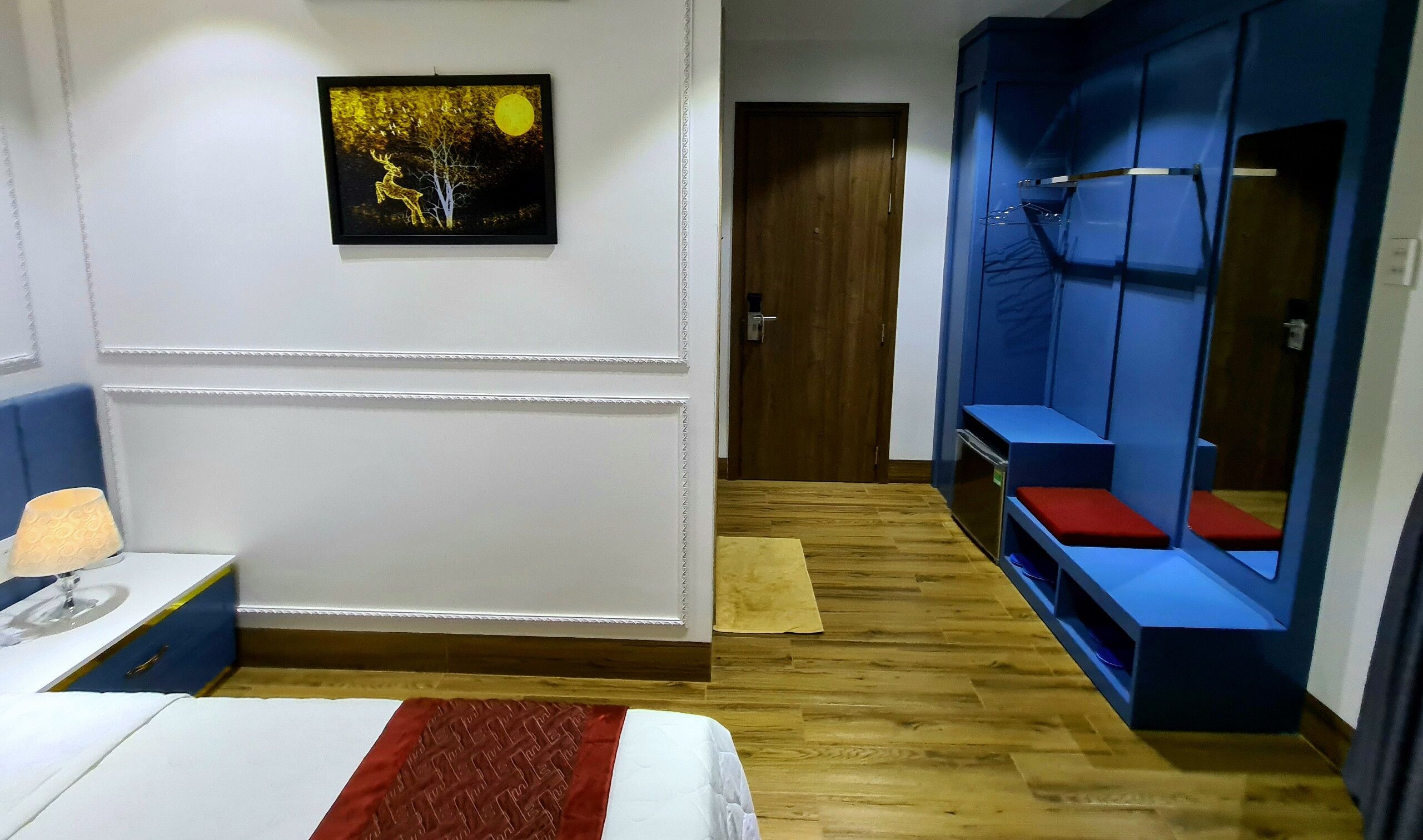 A hotel room with a bed, a bedside table, a lamp, a framed picture on the wall, and a blue wardrobe with a mirror.