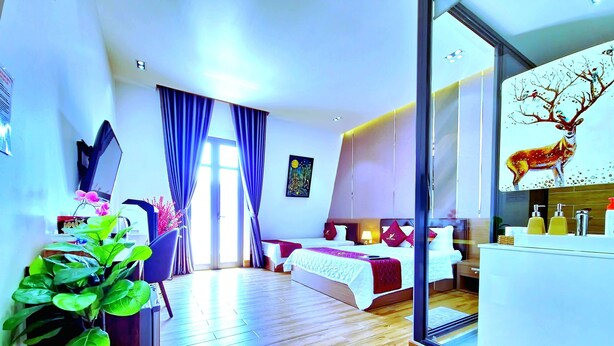 A modern hotel room with a wooden floor, a bed with a red blanket, a desk with a computer, a chair, a potted plant, and a large window with curtains. - Thu Le Hotel (Cao Lanh)