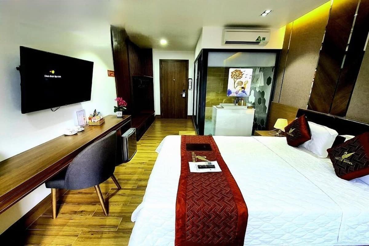 Quarto de hotel com uma cama, uma escrivaninha, uma cadeira, televisão e banheiro visível através de uma porta de vidro.