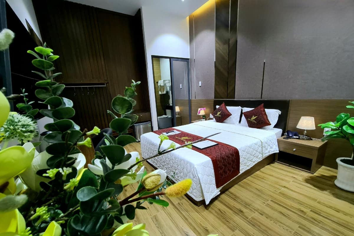 Quarto de hotel moderno com uma cama grande, piso de madeira, mesa de cabeceira com abajur e um espelho.