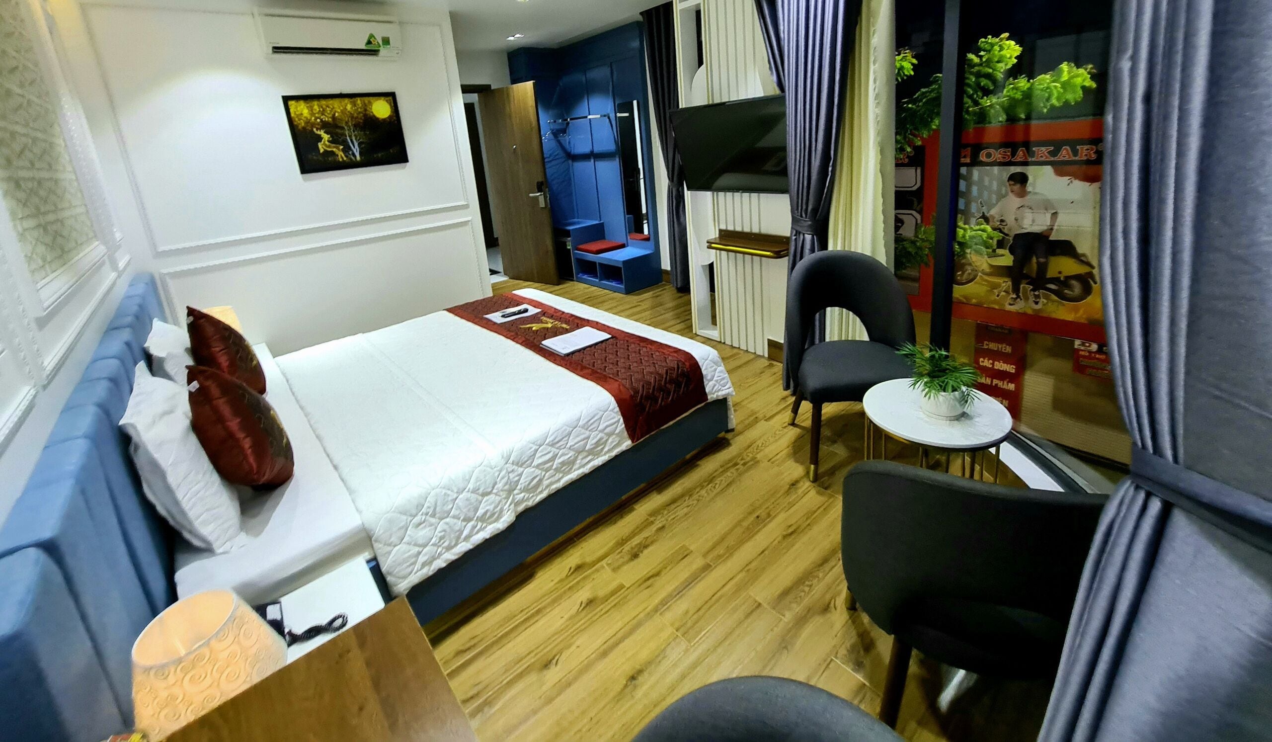 Quarto de hotel com uma cama, uma mesinha com uma planta, uma cadeira, uma escrivaninha e uma janela com cortinas.