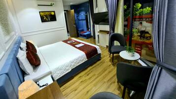 Quarto de hotel com uma cama, uma mesinha com uma planta, uma cadeira, uma escrivaninha e uma janela com cortinas.