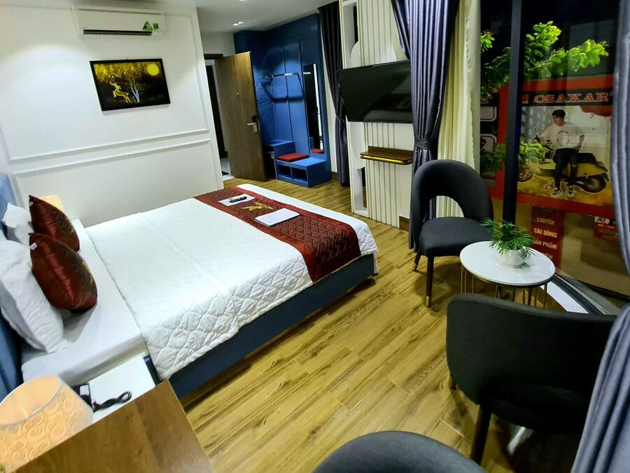 A hotel room with a bed, a small table with a plant, a chair, a desk, and a window with curtains.