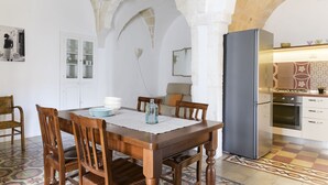 Zona pranzo con tavolo in legno, sedie e un lampadario. Si vede una cucina sullo sfondo con frigorifero e piano cottura.