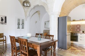 Een eetruimte met een houten tafel, stoelen en een kroonluchter. Achtergrond is een keuken met een koelkast en fornuis.