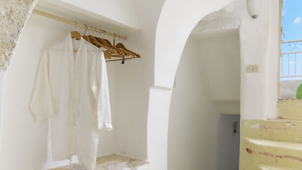 Una stanza dalle pareti bianche, con un appendiabiti in legno che sorregge indumenti bianchi. Una scala con ringhiera gialla conduce a un balcone esterno.