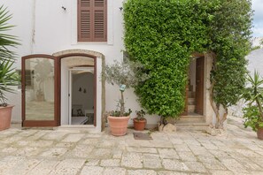 Une cour pavée de pierres, avec un bâtiment blanc, deux portes en bois et des plantes en pot.