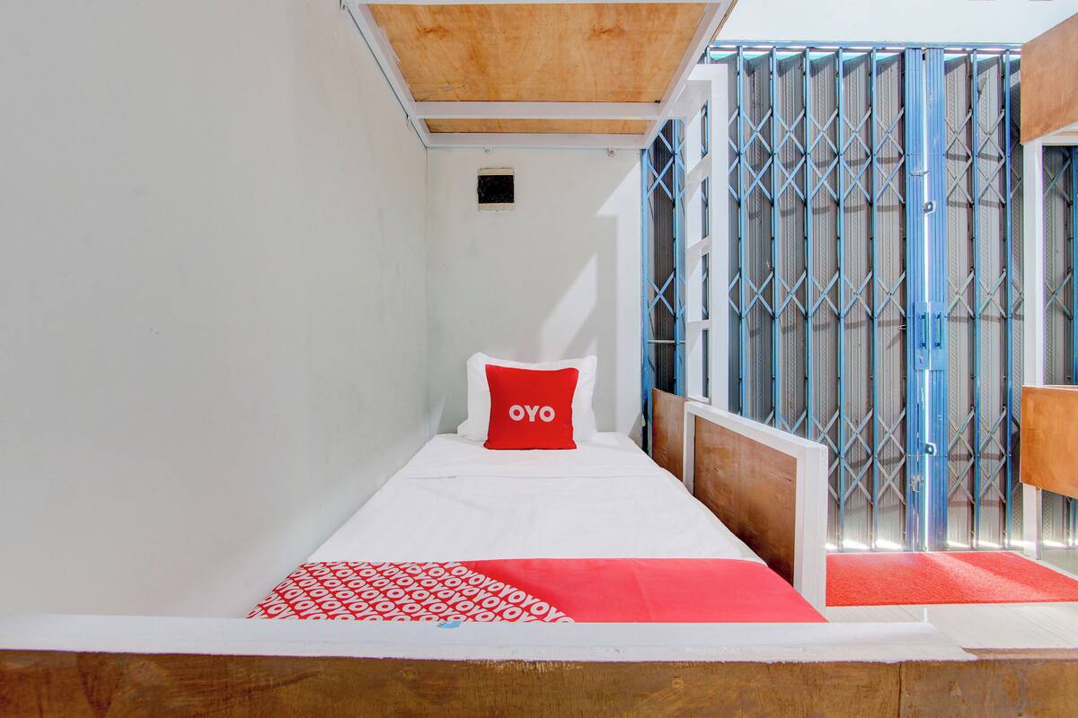 Una cama individual con almohada roja, ropa de cama blanca y una manta estampada en rojo y blanco.