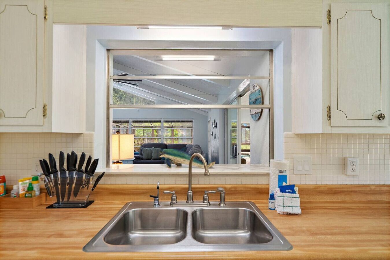 A kitchen with a double sink, a window with a view of a living room, and a tiled backsplash.