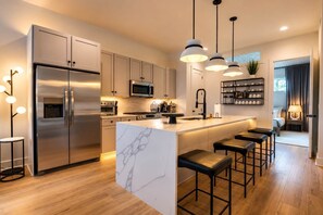 Une cuisine moderne dotée d’un îlot central, d’appareils électroménagers en acier inoxydable et de luminaires suspendus.