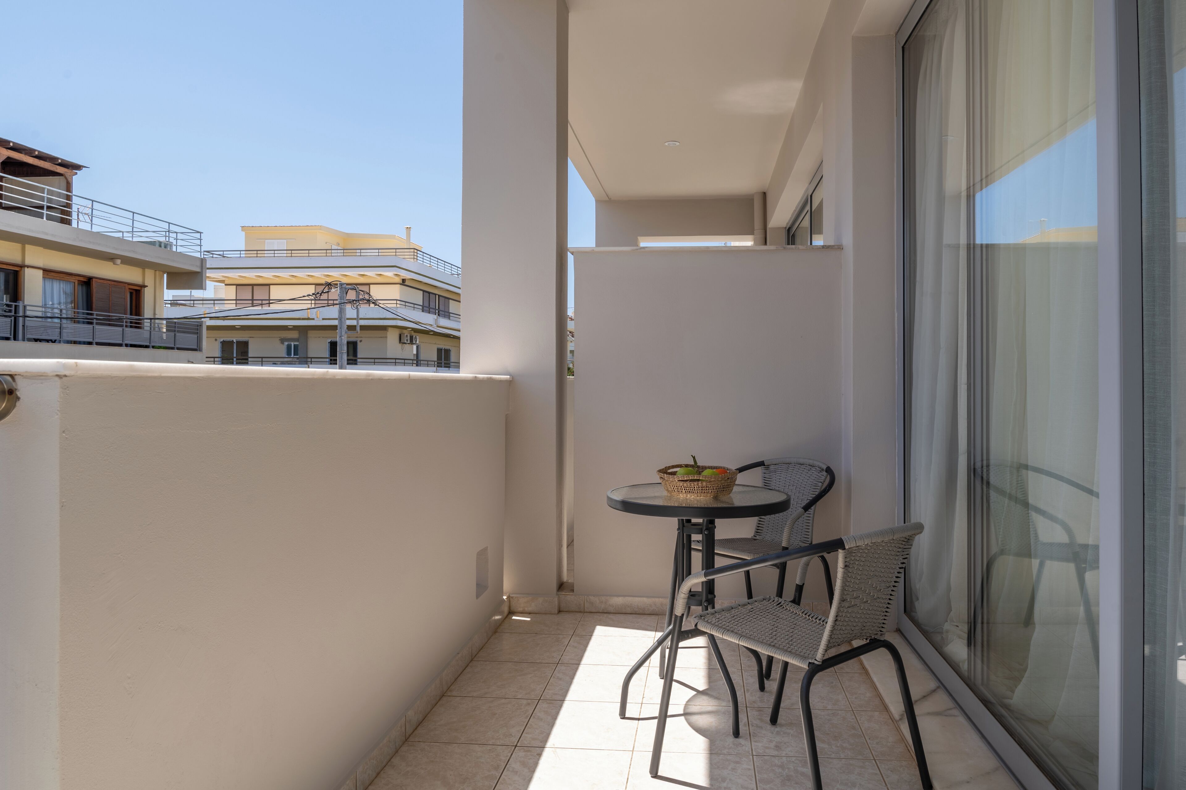 A balcony with a table and two chairs, a window, and a view of neighboring buildings.
