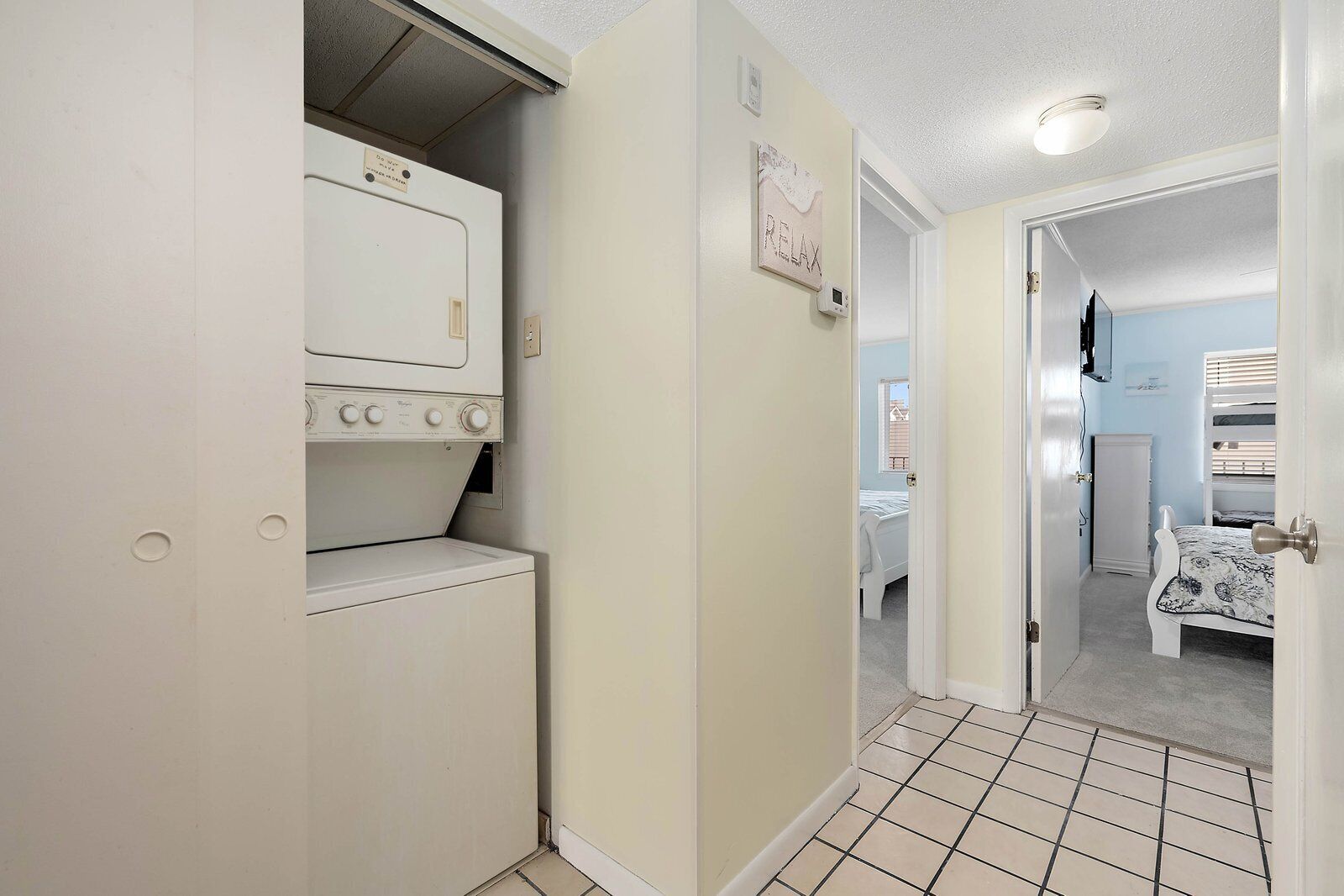 Een wasserette met een wasmachine en droger, een deur naar een slaapkamer met een stapelbed, en een 'RELAX'-bord aan de muur.