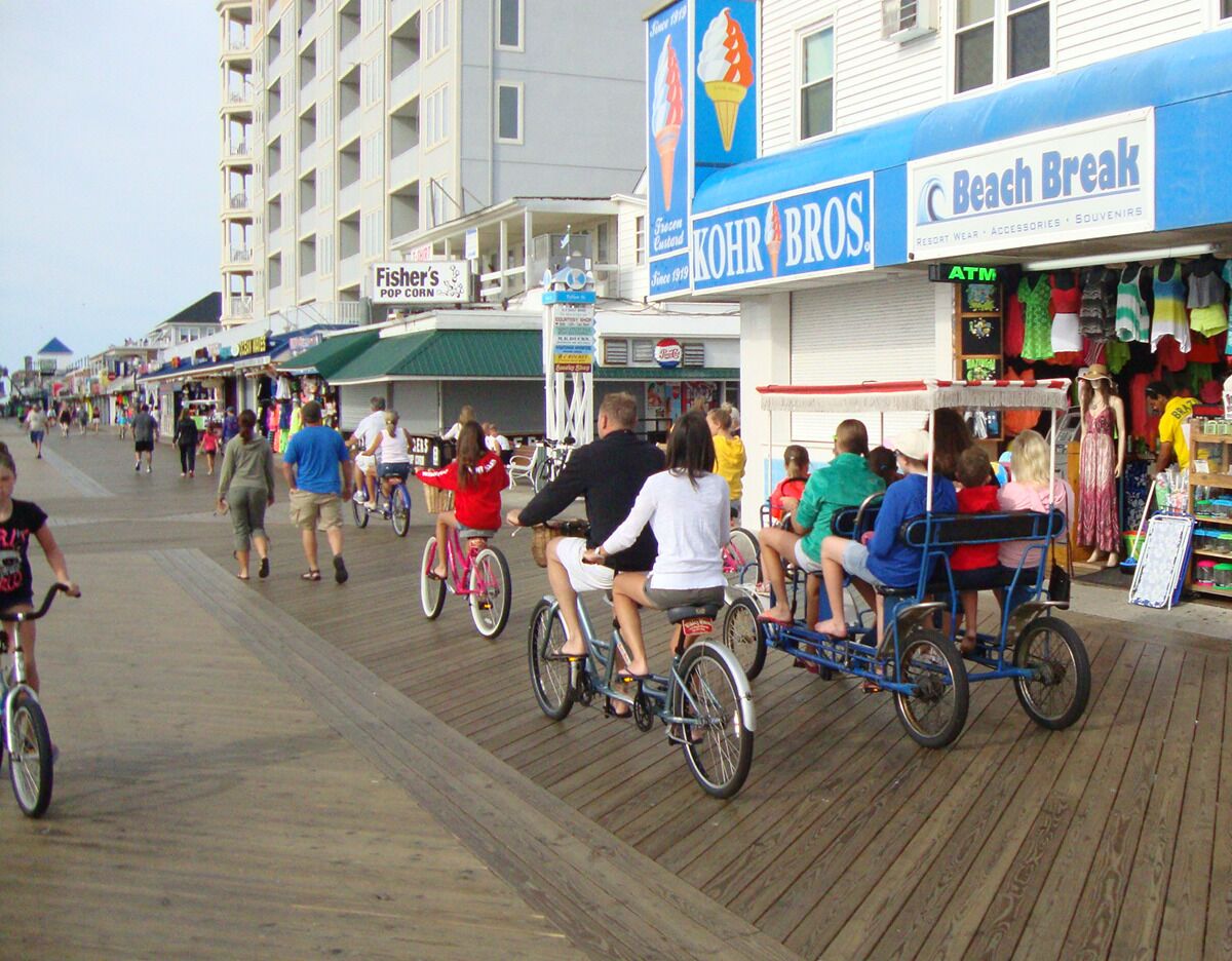 A boardwalk with people riding bicycles and a cart, shops, and a sign for 'Beach Break'.
