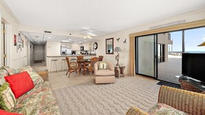 A living room with a sofa, a dining table with chairs, a kitchen area, and a large sliding glass door leading to an outdoor area. - Oceanfront for 9 people at the Rainbow / outdoor pool (Ocean City)