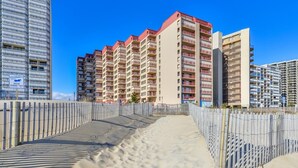 A beachfront hotel with multiple high-rise buildings, a sandy walkway, and a wooden fence. - Oceanfront for 9 people at the Rainbow / outdoor pool (Ocean City)