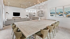 A spacious dining area with a large wooden table and modern chairs, a chandelier, and a kitchen in the background. - Seven Palms Pinnacle|Easy Beach Access|New Construction|Private Pool|Gulf Views (Inlet Beach)