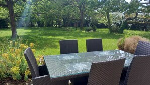 Een terras buiten met een tafel met glazen blad en rieten stoelen, omgeven door een weelderige tuin en bomen.