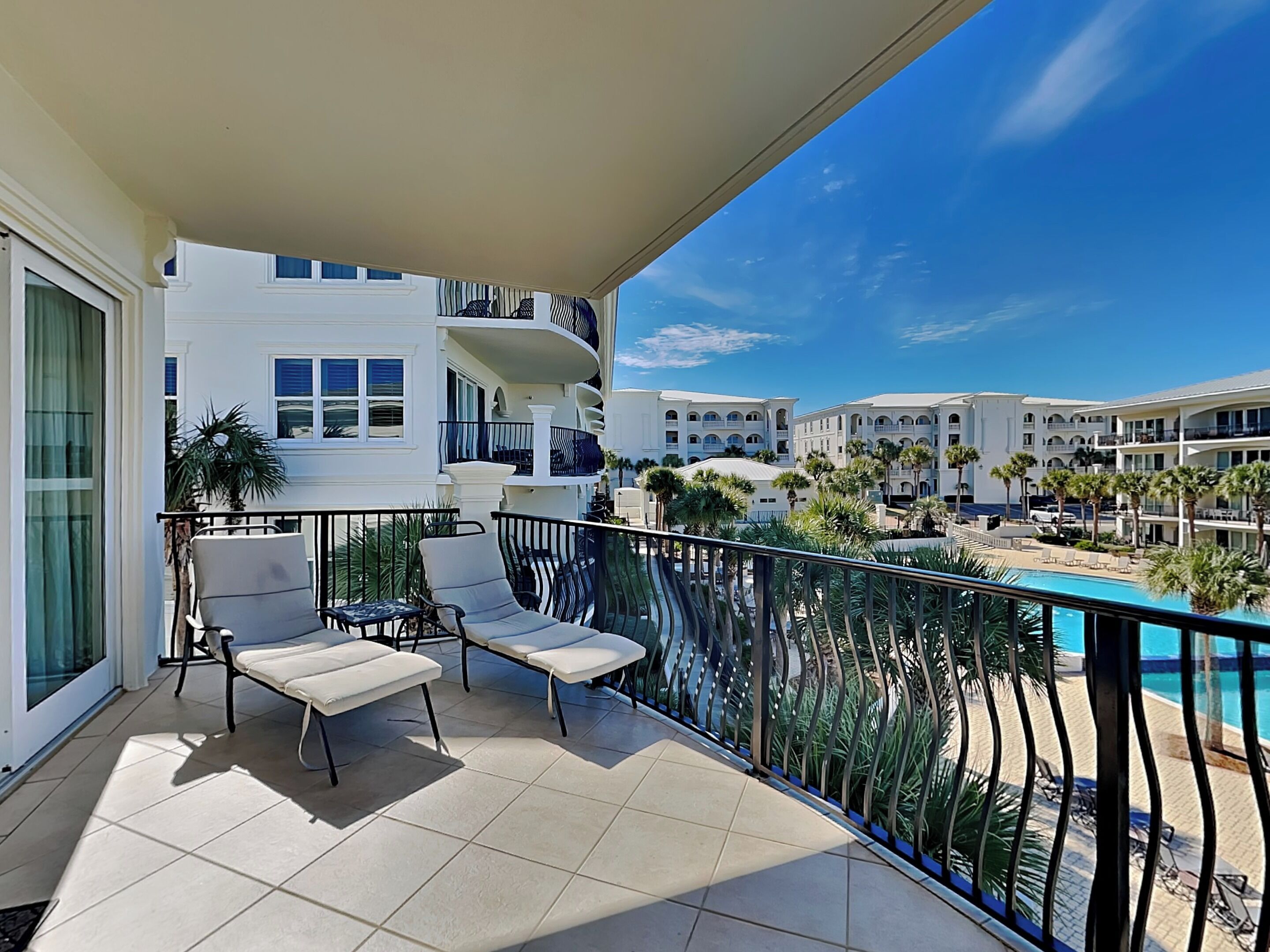 A balcony with two lounge chairs and a small table, overlooking a pool and a resort building.