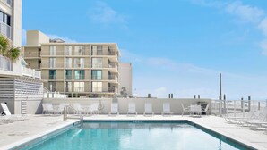 Ein Schwimmbecken mit Liegestühlen, ein Gebäude mit mehreren Balkonen und ein klarer Himmel.