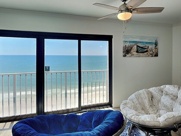 A room with a large sliding glass door leading to a balcony with a view of the ocean. There is a ceiling fan and a painting of a boat on the wall.