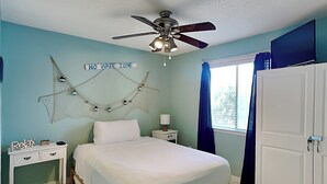 A bedroom with a bed, a nightstand, a ceiling fan, a 'NO WAKE ZONE' sign, and a window with curtains. - Island Home #102 | Close to Beach and Destin Harbor (Destin)