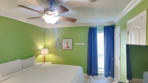 Une chambre d’hôtel avec un lit, un ventilateur de plafond, une table de chevet avec une lampe, un tableau au mur et une fenêtre avec des rideaux.
