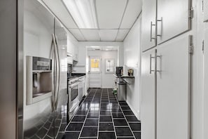 A narrow kitchen with stainless steel appliances, white cabinets, and black tiled flooring.