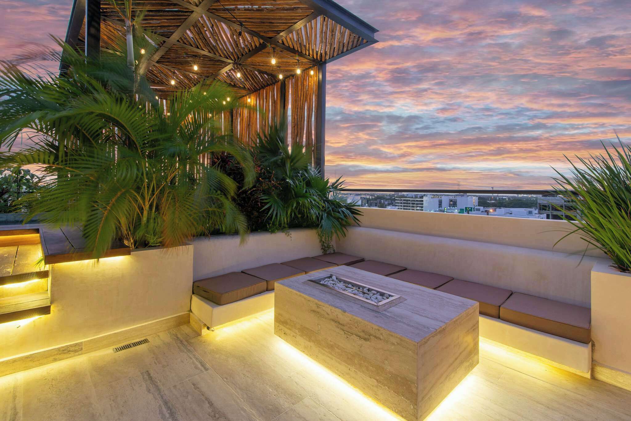 A rooftop terrace with a seating area, plants, and a view of the cityscape at sunset.