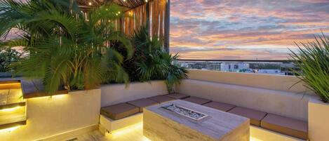 A rooftop terrace with a seating area, plants, and a view of the cityscape at sunset.