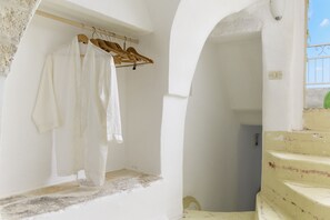 Une pièce aux murs blancs, où un portant en bois supporte des vêtements blancs. Un escalier, dont le mur est d’une couleur jaune pâle, mène à un balcon extérieur.