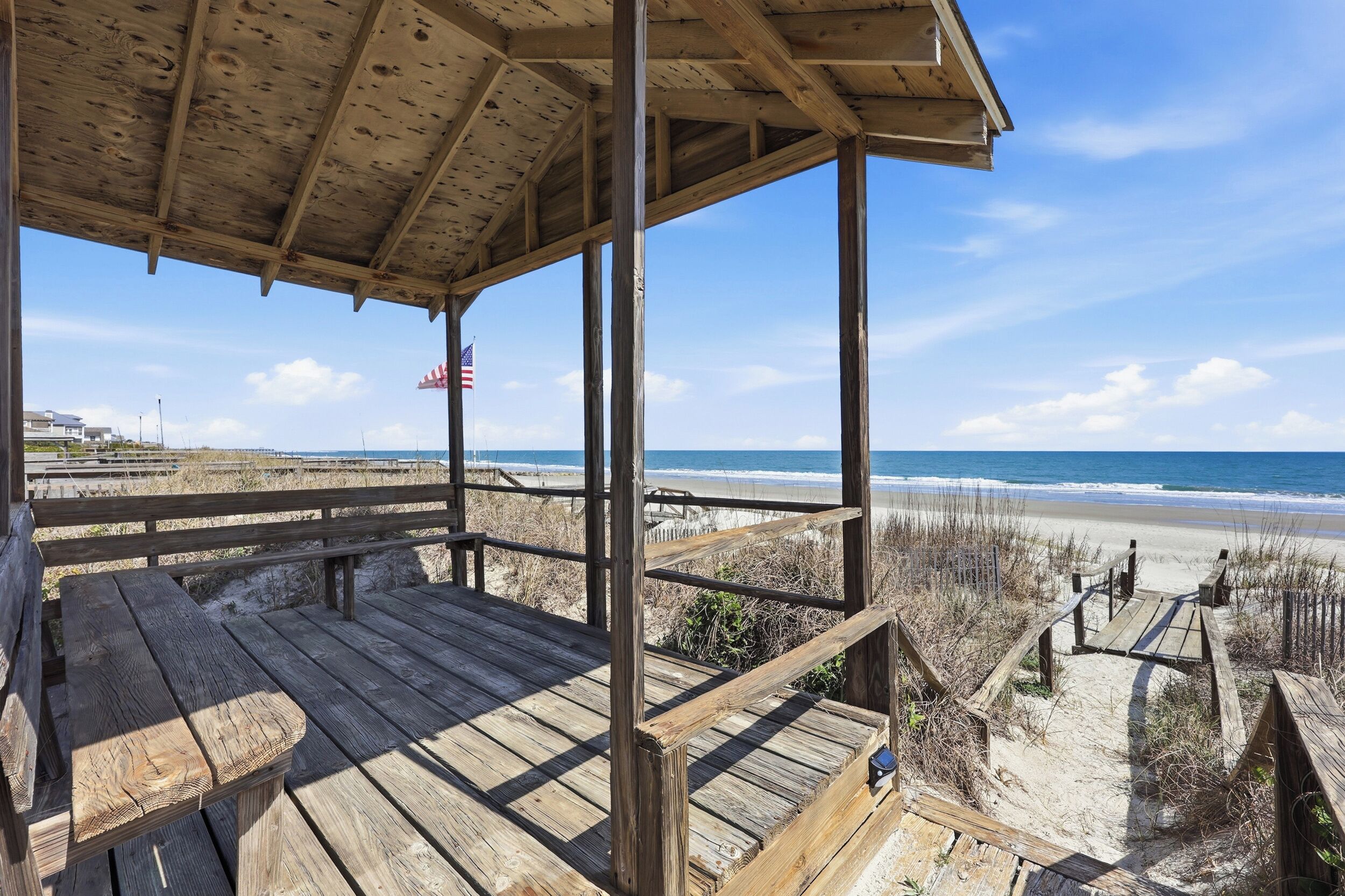 A wooden deck with a view of the beach and ocean, a flag, and a bench.