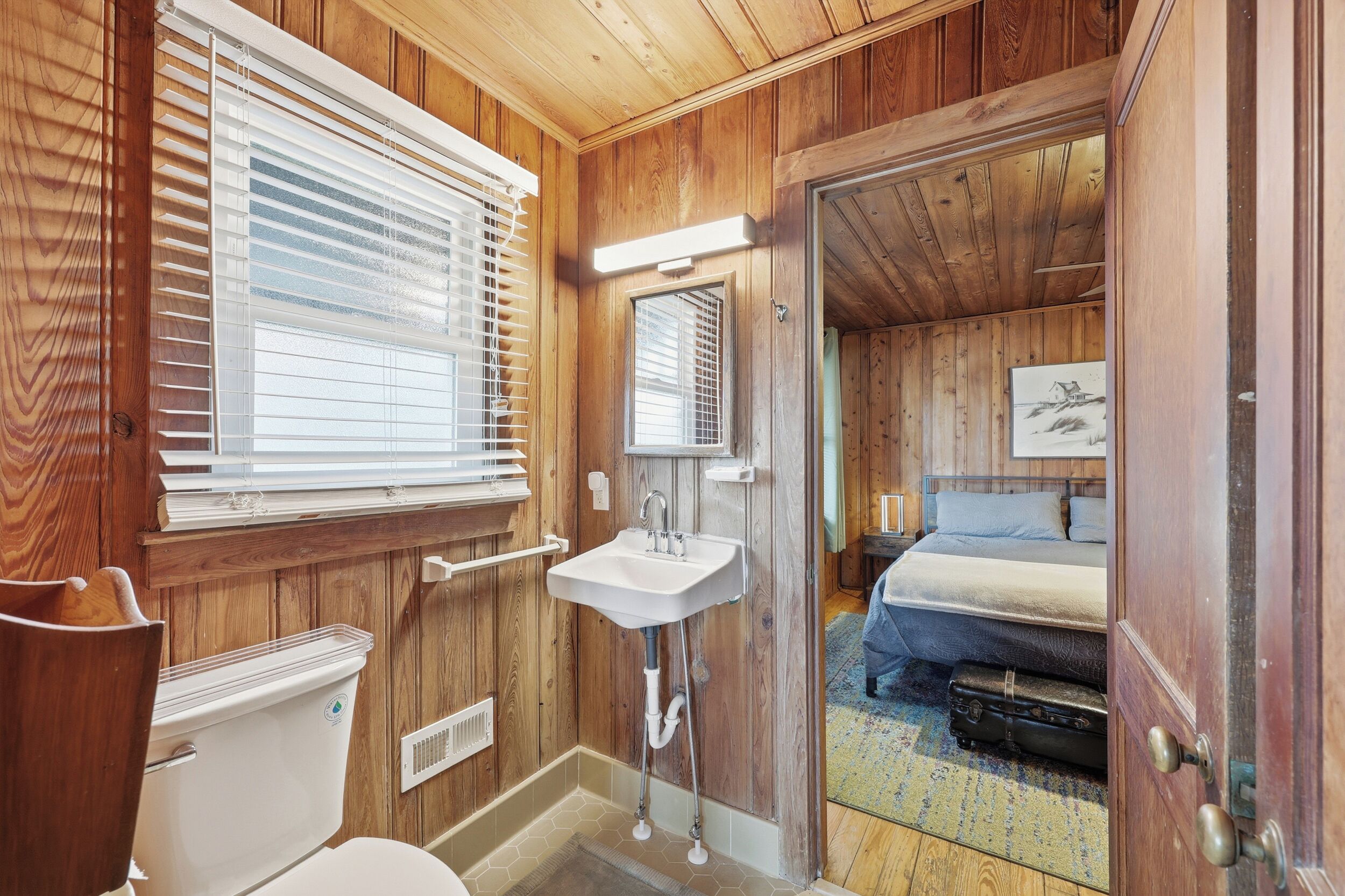 Une salle de bain aux murs lambrissés, équipée de toilettes, d’un lavabo et de stores aux fenêtres.