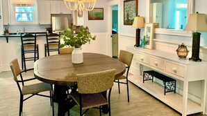 A dining area with a round table, wicker chairs, a chandelier, a white cabinet with a mirror, and a kitchen in the background.