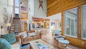 A spacious living area with a wooden ceiling, a kitchen in the background, and a large window with blinds. - Nature Adventure with Access to Exclusive Golf (Sunriver)