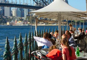 A boat tour with people seated under a canopy, a waterfront view, and a bridge in the background.