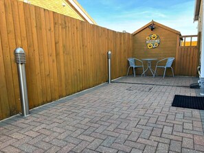 Un espace patio agrémenté d’une clôture en bois, de deux bornes en métal et d’une pancarte « Bienvenue » ornée de tournesols. On y trouve deux chaises en métal et une petite table.