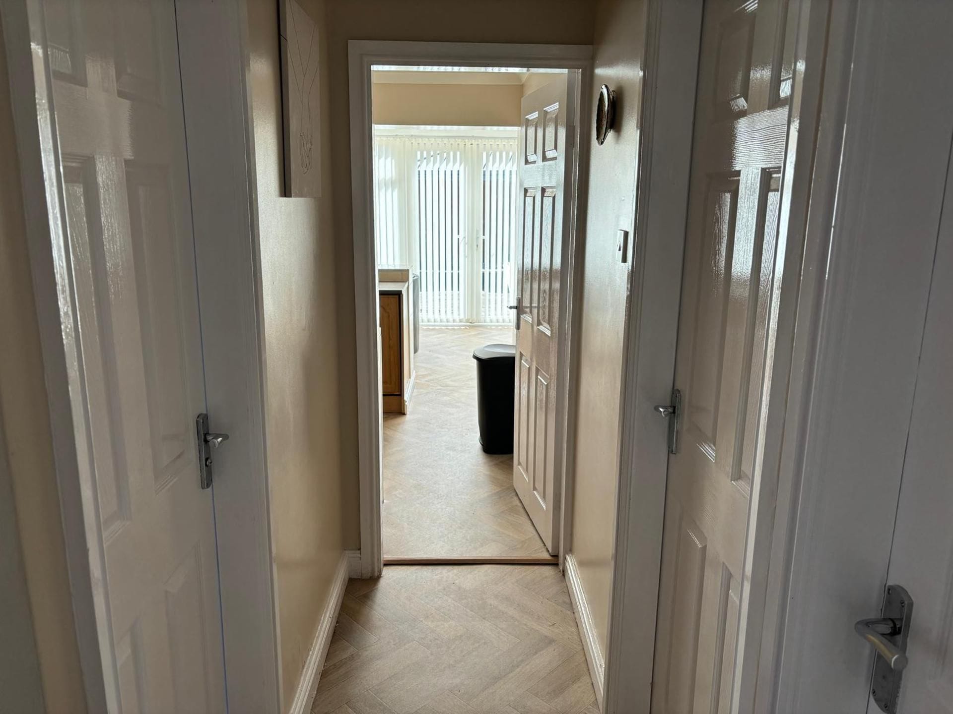A hallway leading to a room with a door, a trash bin, and a window with blinds.