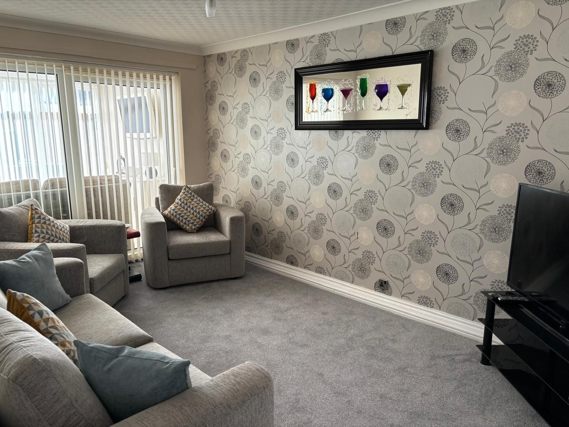 Een woonkamer met een grijze bank, fauteuils, een televisie en een lijst met daarop afbeelding van wijn glazen aan de muur.