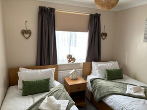 Une chambre avec deux lits, chacun agrémenté d’un oreiller vert et d’une serviette blanche. Une table de chevet en bois se trouve entre les lits, et il y a une fenêtre avec des rideaux.