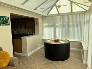A modern kitchen with wooden cabinets, a dining area with a round table, and a large window with blinds. - 2-bed Bungalow, Sleeps 6, Parking & Pet Friendly (Skegness)